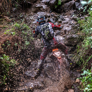Enduro racer climbing a very steep, wet, & rocky hillside. His Plews Tyre rear tire is just throwing dirty water