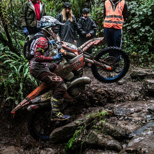 Enduro racer pulling a wheelie up a muddy and rocky terrain as he passes a few racing spectators. His bike is running Plews Tyres.