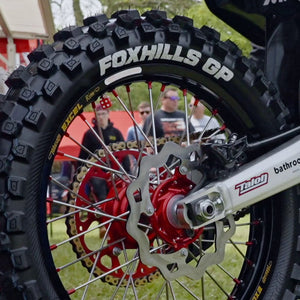 Close up of the rear tire and swing arm of a motocross bike in an outdoor grass setting. Whitewall lettering of FOXHILLS GP and Plews Tyres prominently displayed.