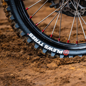 Super close up on a rear dirt bike tire with the Plews Tyres logo prominently displayed in white against the black rubber.