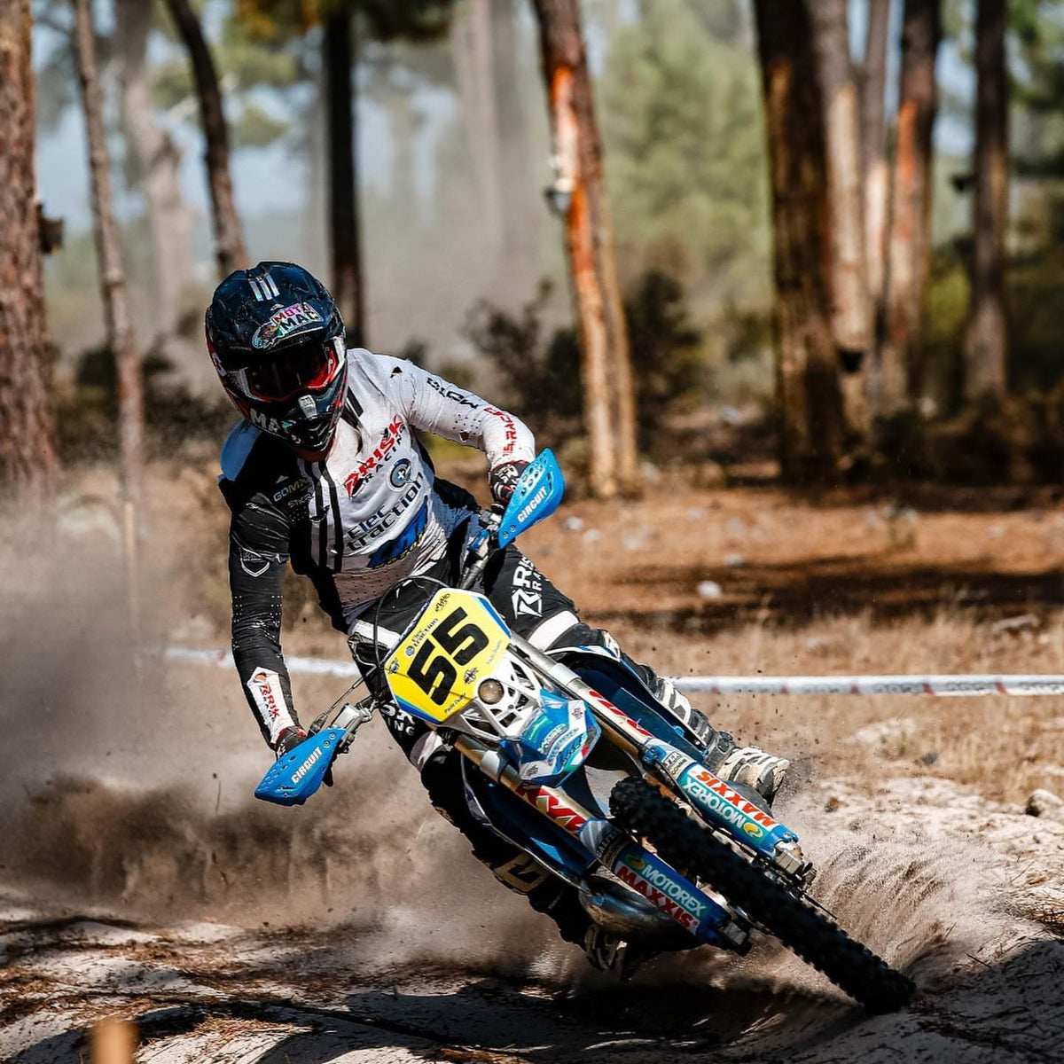 Motocross rider with number 55 leans into a fast sandy turn on a dirt trail through a pine forest during an off-road enduro race, kicking up a cloud of dust.
