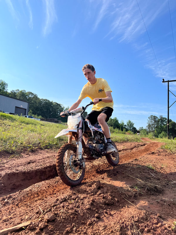 male riding a pit bike on a track wearing a yellow 'spark wings' Risk Racing t-shirt