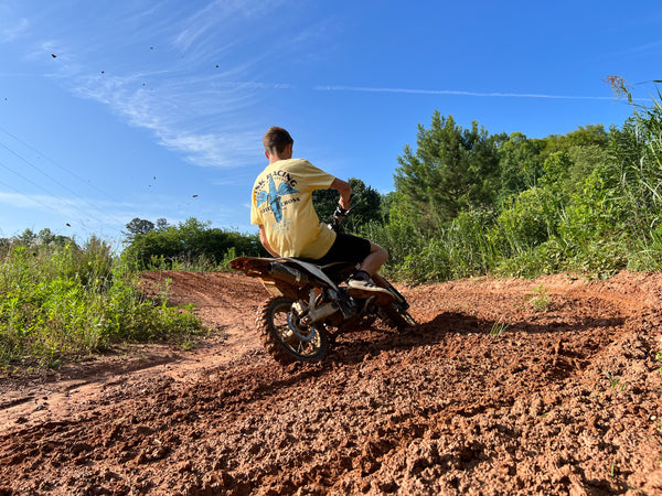 male riding a pit bike on a track wearing a yellow 'spark wings' Risk Racing t-shirt