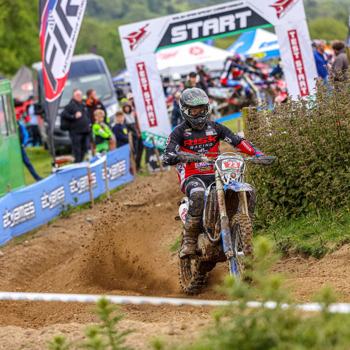 Motocross rider number 23 wearing Risk Racing gear powers through a sandy section at the start of an enduro race, with spectators, flags, and the official start arch visible in the background.