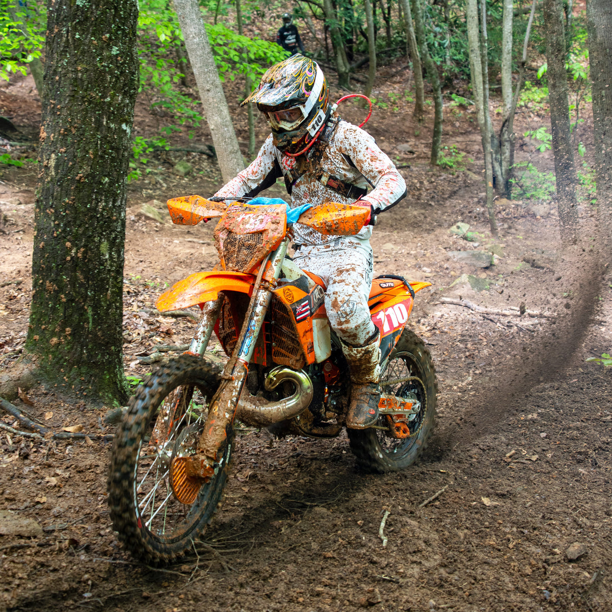 Off-road rider wearing white gear and helmet number 110 powers through a muddy forest trail on an orange dirt bike, flinging dirt as they navigate tight terrain surrounded by trees.