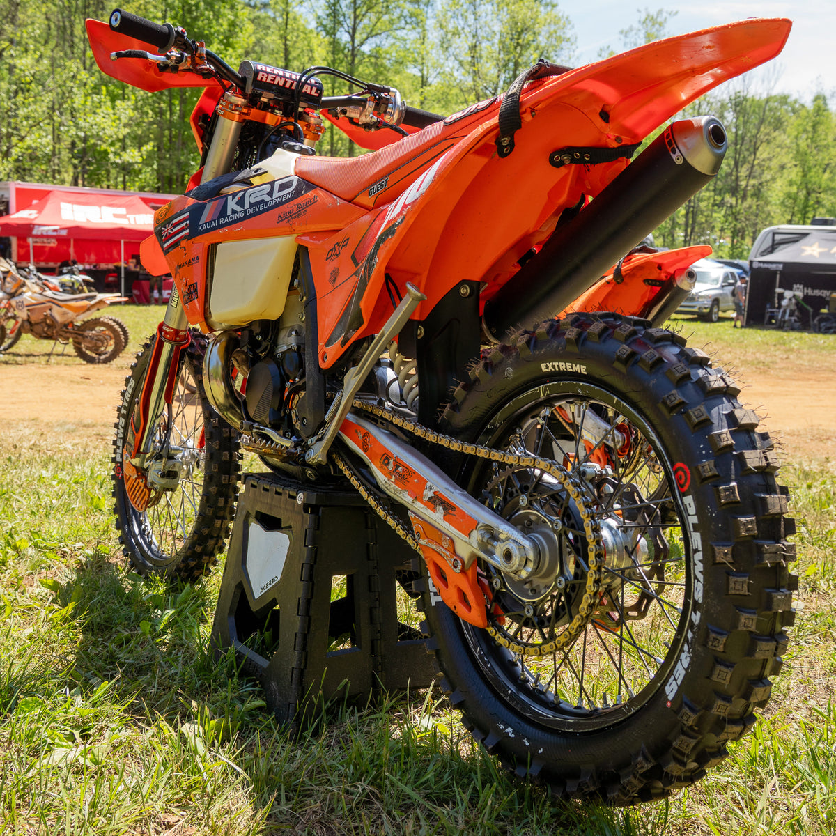 Bright orange KTM dirt bike with KRD and Plews Tyres branding displayed on a black stand at an outdoor motocross event, with detailed view of the rear suspension, tire tread, and chain assembly.