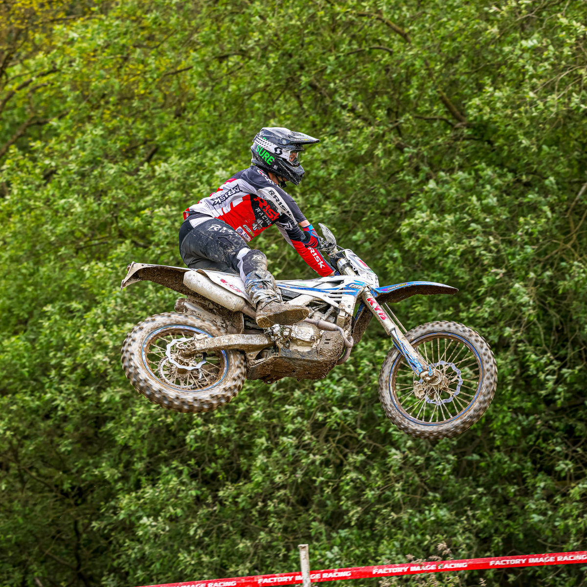 Motocross rider in red and black Risk Racing gear catches air during a jump at an off-road enduro event, with dense green trees and Factory Image Racing tape in the background.