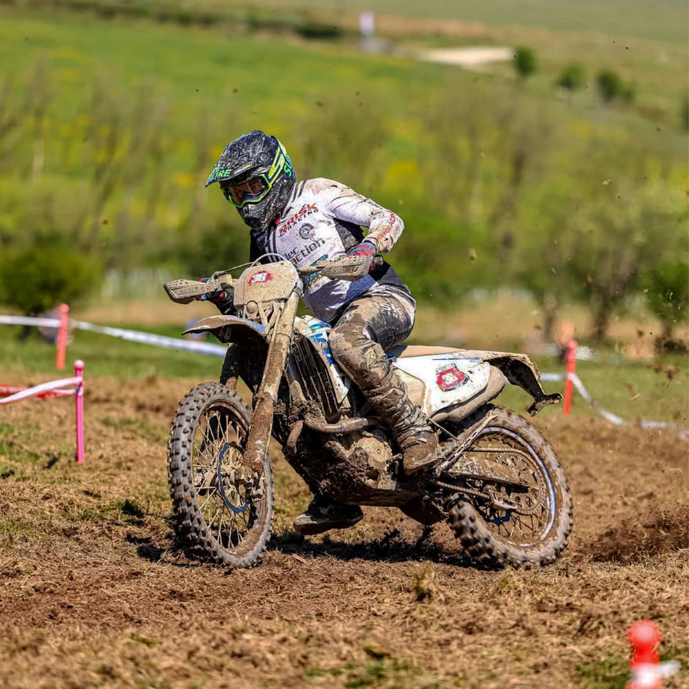 Motocross rider speeds through a muddy section of a grassy enduro racecourse, leaning into a turn with dirt flying, surrounded by ribbon markers and open countryside in the background.