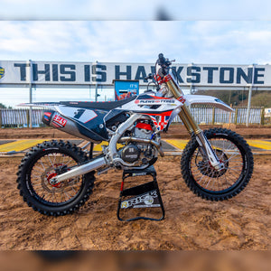 motocross bike wearing Plews tires sitting on a Risk Racing A.T.S. stand at an MX track with a giant sign in the background that reads: this is hawkstone